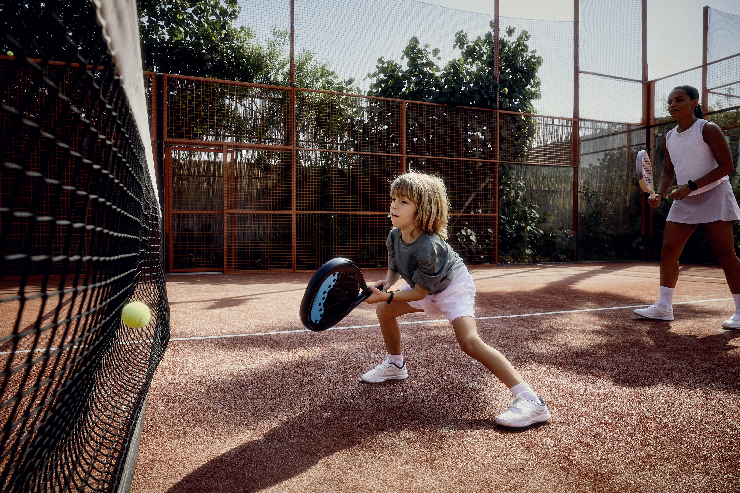 A child and a woman play padel on a clay court.